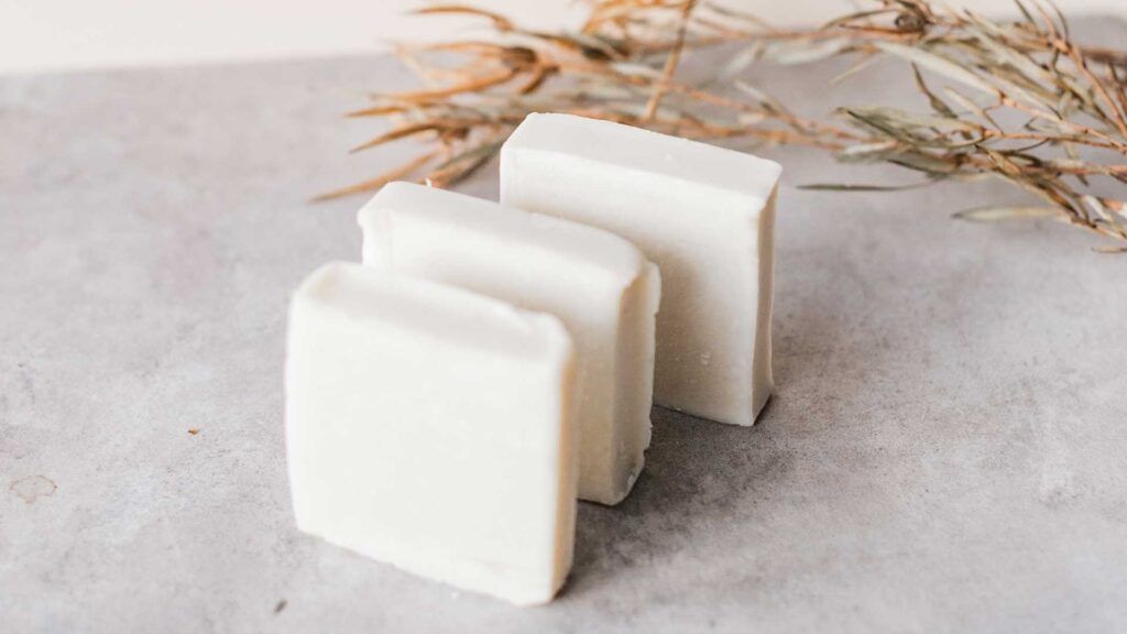 Tres pastillas de jabón blanco natural sobre una superficie de piedra con hojas secas decorativas al fondo.