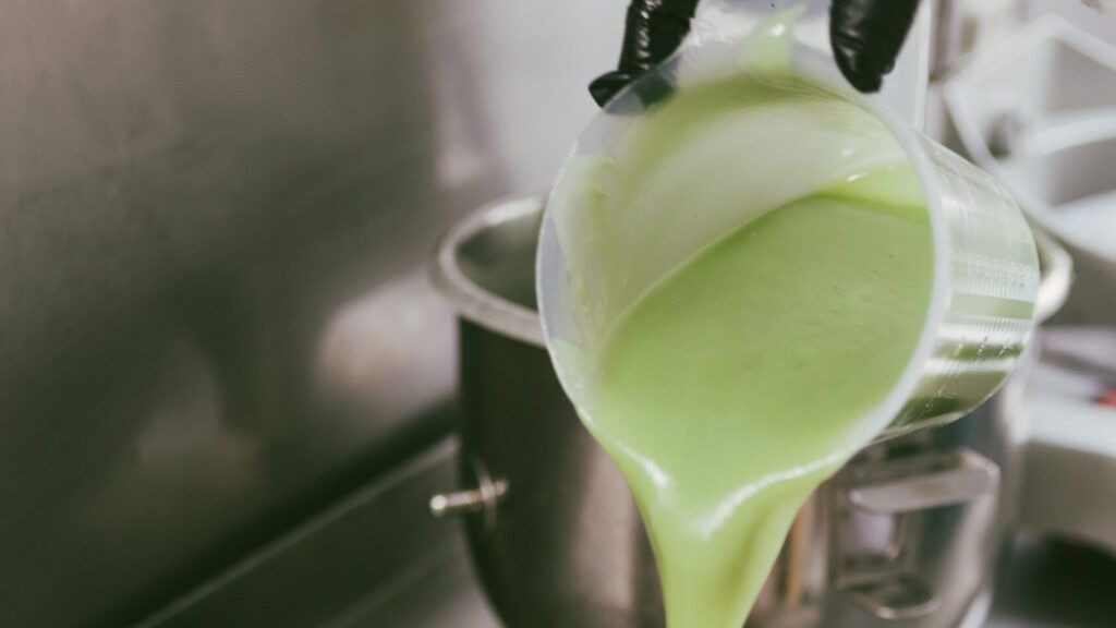 Mezcla líquida verde siendo vertida desde un recipiente transparente a una olla metálica durante el proceso de fabricación de jabón artesanal.