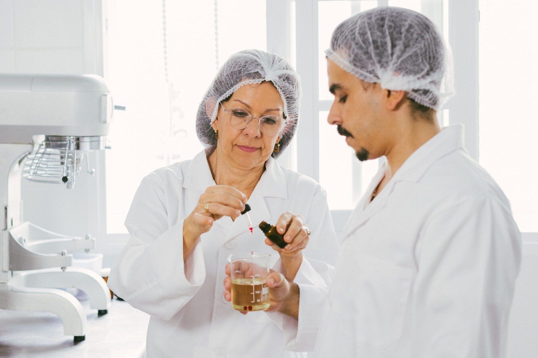 Dos personas con bata y gorro de laboratorio trabajando con un gotero y vaso medidor en un entorno de formulación cosmética.