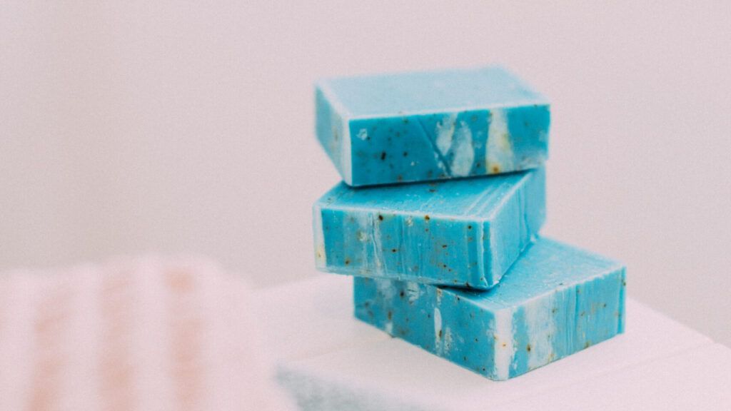 Tres barras de jabón artesanal azul con partículas exfoliantes apiladas sobre una superficie blanca.