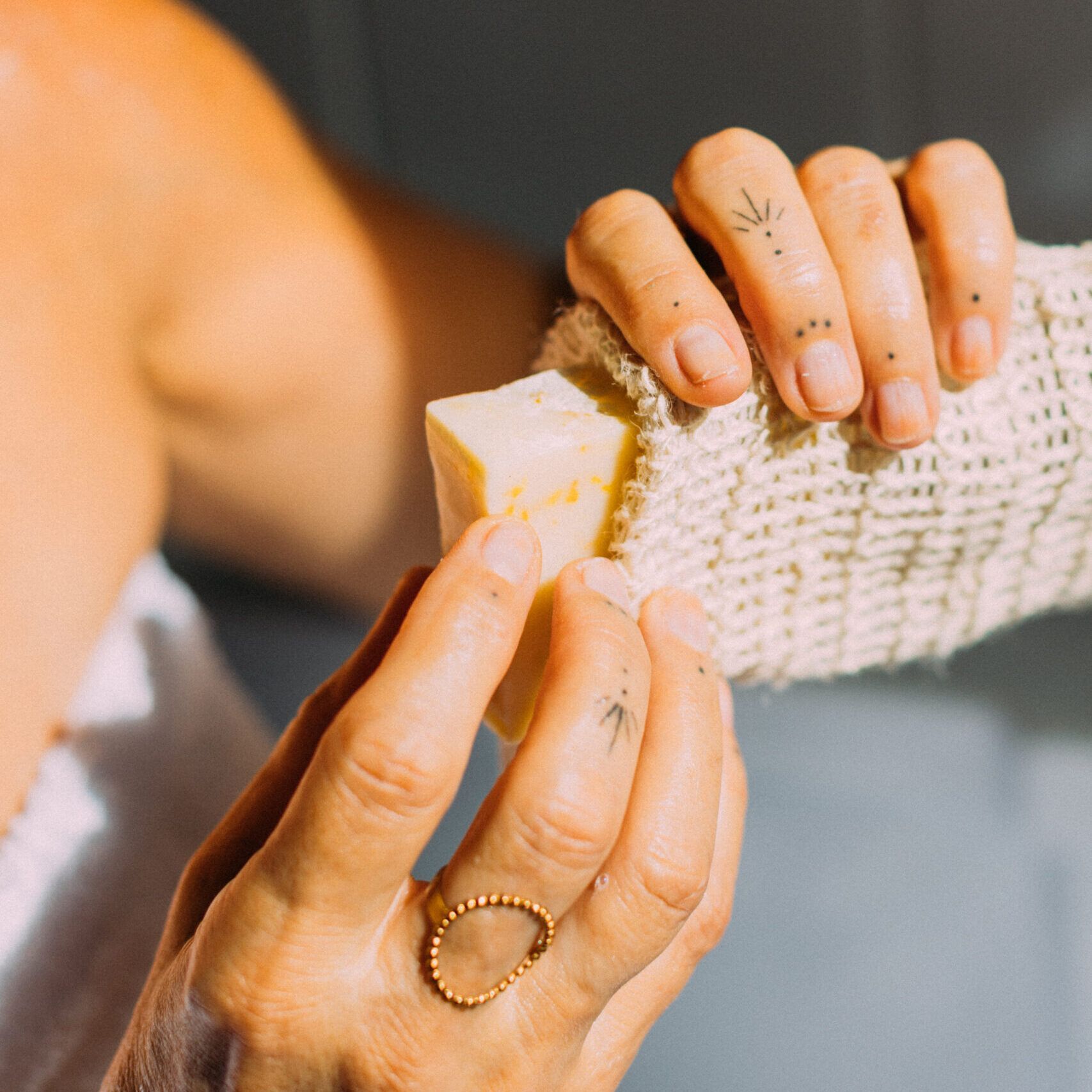 Persona insertando un jabón artesanal con pétalos de caléndula en una bolsita exfoliante de fibra natural.