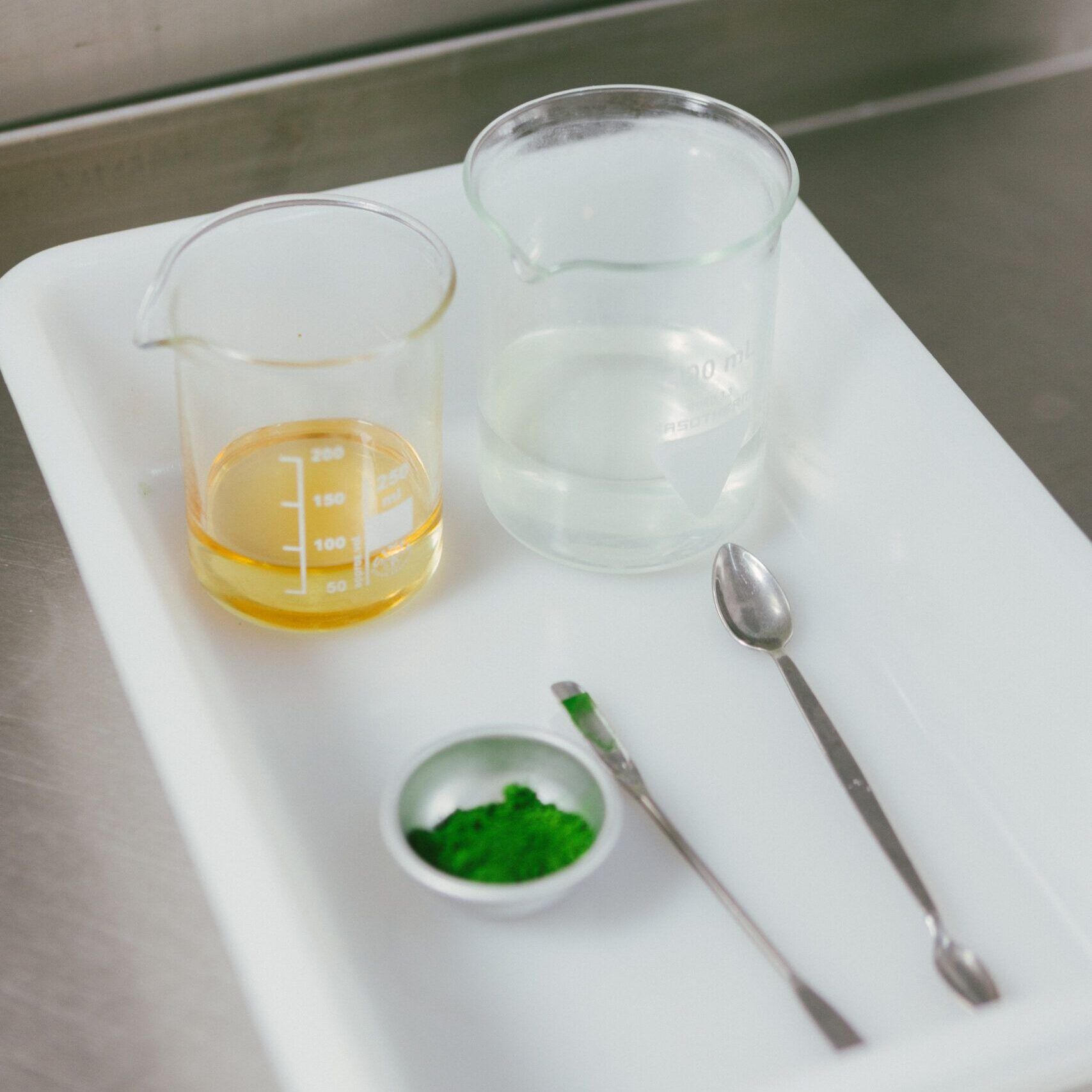 Bandeja blanca con utensilios de laboratorio: vasos medidores con líquidos, cucharas metálicas y polvo verde en un cuenco.